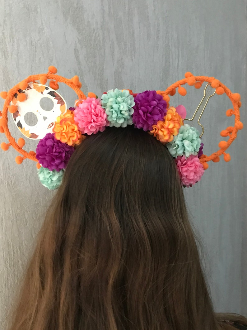 K&ouml;nnte beinhalten: Ein orangefarbenes Stirnband mit einem Totenkopfdesign und bunten Bommeln. Das Stirnband ist mit verschiedenen Blumen in den Farben Rosa, Lila, Orange und Gr&uuml;n verziert.