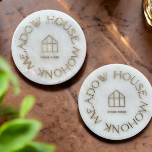Peut inclure: Deux sous-verres ronds blancs avec un lettrage doré qui dit "Stone Ridge Meadow House" en cercle autour d'une icône de maison.