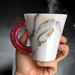 May include: White ceramic mug with a marbled design and a gold letter "C" on the front. The mug has a red handle.