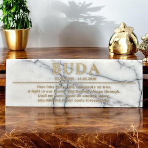 Può includere: Placca commemorativa in marmo bianco con lettere dorate. La targa recita "BUDA 28.07.2020 - 14.02.2025 Il tuo amore è stato un dono, un tesoro così vero. Una luce nei nostri cuori che brilla per sempre. Finché non ci incontreremo di nuovo su un'altra riva, vivrai nelle nostre anime per sempre."