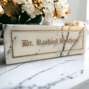May include: A white marble rectangular box with gold trim and the inscription "Dr. Rachael Brothers" in gold lettering.