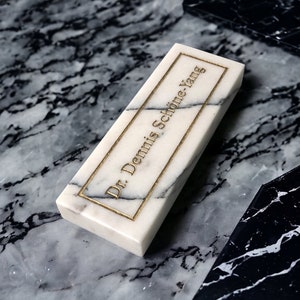 May include: A white marble rectangular plaque with gold lettering that reads "Dr. Dennis Schone Yang". The plaque is sitting on a black and white marble surface.