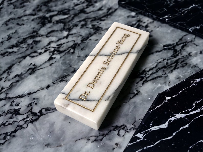 May include: A white marble rectangular plaque with gold lettering that reads "Dr. Dennis Schone Yang". The plaque is sitting on a black and white marble surface.