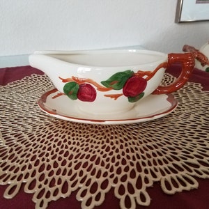 May include: White ceramic gravy boat with a red handle and a brown and white saucer. The gravy boat is decorated with a red apple and green leaves on a brown branch.