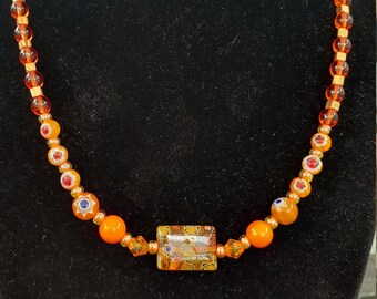 May include: A necklace made of orange and gold beads with a rectangular, multi-colored glass pendant.