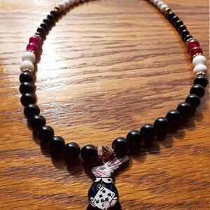 May include: Black beaded necklace with a white and red bead pattern. The necklace has a charm of a white rabbit holding a playing card with the four of spades.