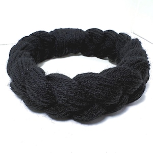 Peut inclure: Un bandeau circulaire fabriqué à partir de fil noir épais tressé en un motif continu et volumineux. L'accessoire présente un aspect doux et texturé avec une couleur sombre uniforme sur toute la structure tressée.