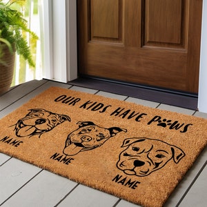May include: A rectangular coir doormat with a brown background. The mat features three dog illustrations and the text "OUR KIDS HAVE PAWS". Below each dog illustration is the word "NAME".
