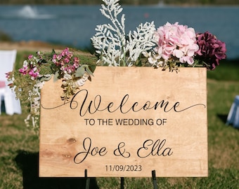 Puede incluir: Un cartel de bienvenida de madera para una boda con el texto "Welcome to the wedding of Joe & Ella 11/09/2023". El cartel está decorado con flores rosas y blancas.
