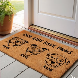 May include: A rectangular doormat made of natural coir fiber. The mat features three stylized dog faces with the text "OUR KIDS HAVE PAWS" and spaces for names. The design is in black against the brown background.
