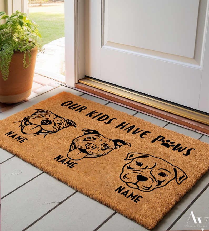 May include: A rectangular doormat made of natural coir fiber. The mat features three stylized dog faces with the text "OUR KIDS HAVE PAWS" and spaces for names. The design is in black against the brown background.