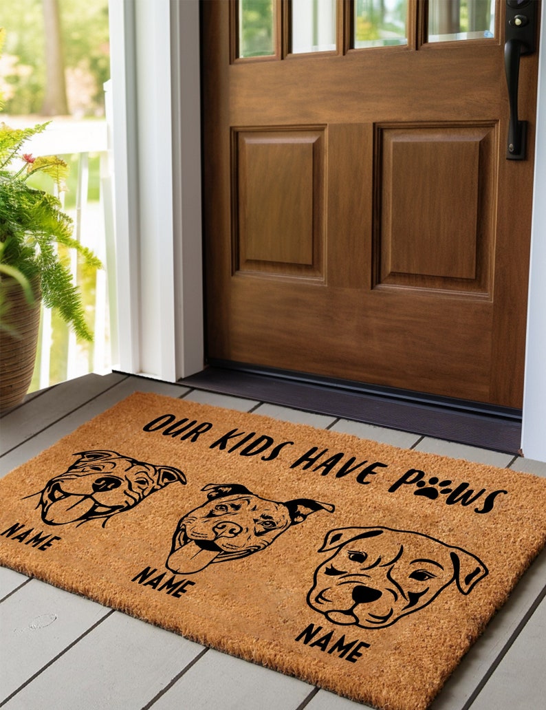 May include: A rectangular coir doormat with a brown background. The mat features three dog illustrations and the text "OUR KIDS HAVE PAWS". Below each dog illustration is the word "NAME".