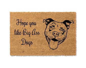 May include: A rectangular doormat with a light brown, textured surface. The mat features the text "Hope you like Big Ass Dogs" in black script, along with a black illustration of a smiling dog's face. The dog has its tongue hanging out.