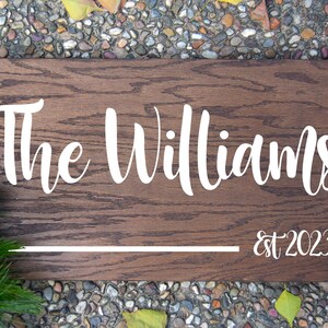Puede incluir: Un letrero rectangular de madera con el texto "The Williams" en escritura blanca, con "est 2023" debajo. El letrero tiene un acabado de vetas de madera marr&oacute;n oscuro y una l&iacute;nea horizontal blanca. Follaje verde en la esquina inferior izquierda.