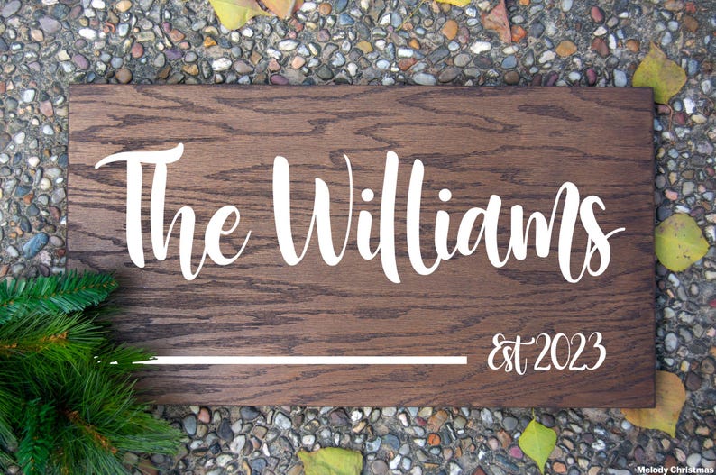 Puede incluir: Un letrero rectangular de madera con el texto "The Williams" en escritura blanca, con "est 2023" debajo. El letrero tiene un acabado de vetas de madera marr&oacute;n oscuro y una l&iacute;nea horizontal blanca. Follaje verde en la esquina inferior izquierda.