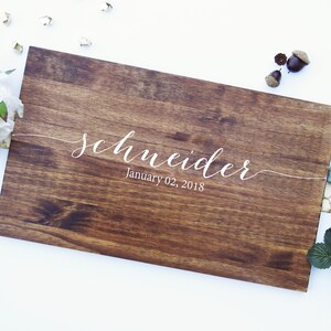 Puede incluir: Un letrero rectangular de madera con el nombre "Schneider" en elegante escritura blanca, junto con la fecha "02 de enero de 2018". El letrero tiene una rica veta de madera marr&oacute;n oscuro y est&aacute; rodeado de elementos decorativos.