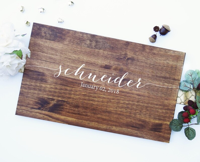 Puede incluir: Un letrero rectangular de madera con el nombre "Schneider" en elegante escritura blanca, junto con la fecha "02 de enero de 2018". El letrero tiene una rica veta de madera marr&oacute;n oscuro y est&aacute; rodeado de elementos decorativos.