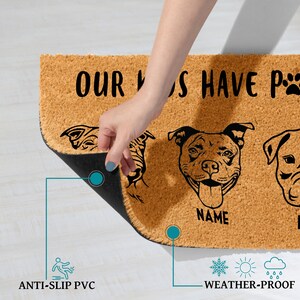 May include: A rectangular doormat made of natural coir fiber, featuring black dog illustrations and the text "OUR DOGS HAVE PAWS". The mat has an anti-slip PVC backing and is weather-proof. A hand is lifting the edge of the mat.