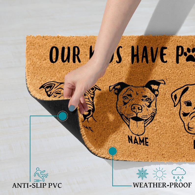 May include: A rectangular doormat made of natural coir fiber, featuring black dog illustrations and the text "OUR DOGS HAVE PAWS". The mat has an anti-slip PVC backing and is weather-proof. A hand is lifting the edge of the mat.