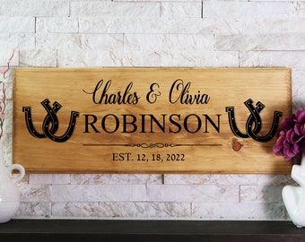 Puede incluir: Un letrero rectangular de madera con acabado en marrón claro natural que presenta texto negro que dice Charles & Olivia ROBINSON y EST. 12, 18, 2022. El diseño incluye dos iconos de herradura negros a cada lado del apellido y una línea.