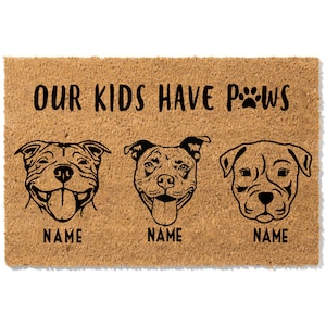 May include: A rectangular doormat made of natural coir fiber. The mat features three stylized dog portraits with the text "OUR KIDS HAVE PAWS" and paw prints. Each dog portrait has the word "NAME" below it.