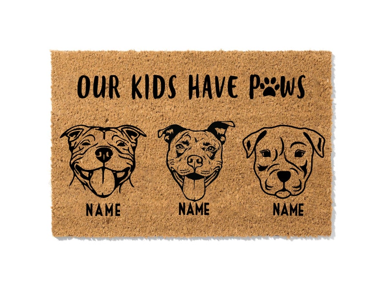 May include: A rectangular doormat made of natural coir fiber. The mat features three stylized dog portraits with the text "OUR KIDS HAVE PAWS" and paw prints. Each dog portrait has the word "NAME" below it.