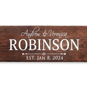 Puede incluir: Un letrero rectangular de madera con tinte marr&oacute;n oscuro. El letrero presenta los nombres "Andrew & Veronica ROBINSON" en escritura blanca y letras de bloque, con "EST. JAN 8, 2024" debajo. El fondo es blanco.
