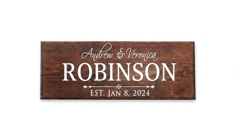 Puede incluir: Un letrero rectangular de madera con tinte marr&oacute;n oscuro. El letrero presenta los nombres "Andrew & Veronica ROBINSON" en escritura blanca y letras de bloque, con "EST. JAN 8, 2024" debajo. El fondo es blanco.