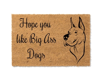 May include: A rectangular doormat with a light brown, textured surface. The mat features the text "Hope you like Big Ass Dogs" in black script, along with a black line drawing of a dog's head.
