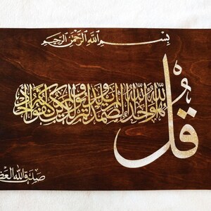 Puede incluir: Placa de madera con caligraf&iacute;a isl&aacute;mica dorada y blanca. El fondo de vetas de madera marr&oacute;n oscuro contrasta con la elegante escritura. El texto est&aacute; en &aacute;rabe, con un estilo art&iacute;stico y fluido. Ideal para la decoraci&oacute;n del hogar.