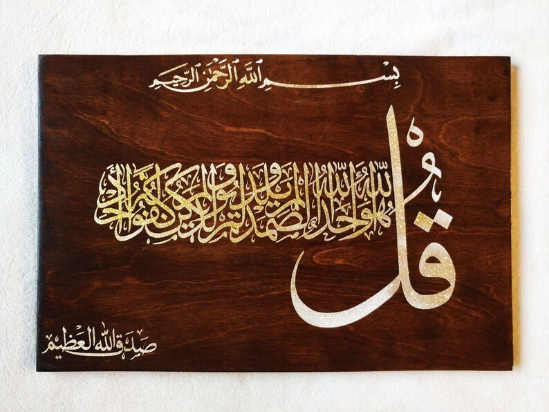Puede incluir: Placa de madera con caligraf&iacute;a isl&aacute;mica dorada y blanca. El fondo de vetas de madera marr&oacute;n oscuro contrasta con la elegante escritura. El texto est&aacute; en &aacute;rabe, con un estilo art&iacute;stico y fluido. Ideal para la decoraci&oacute;n del hogar.