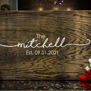 Puede incluir: Un letrero de madera con el nombre "mitchell" en fuente cursiva, con la palabra "The" arriba y "Est. 09.01.2021" abajo. La madera tiene un veteado marr&oacute;n oscuro y texturizado. Un peque&ntilde;o arreglo de flores rojas y blancas est&aacute; en la esquina inferior derecha.