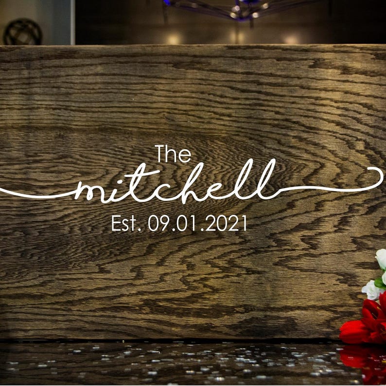 Puede incluir: Un letrero de madera con el nombre "mitchell" en fuente cursiva, con la palabra "The" arriba y "Est. 09.01.2021" abajo. La madera tiene un veteado marr&oacute;n oscuro y texturizado. Un peque&ntilde;o arreglo de flores rojas y blancas est&aacute; en la esquina inferior derecha.