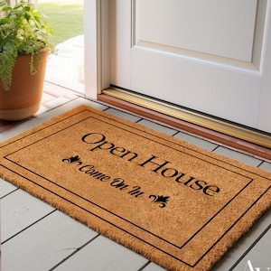 K&ouml;nnte beinhalten: Eine braune Kokosmatte mit schwarzem Rand und dem Text "Open House Come On In" in schwarzen Buchstaben.