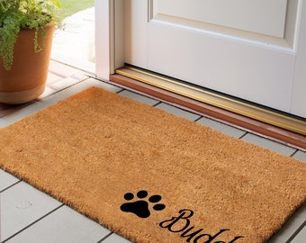 May include: A rectangular doormat made of natural brown coir fiber. The mat features a black paw print and the name "Buddy" in a cursive font. The mat is placed in front of a white door.