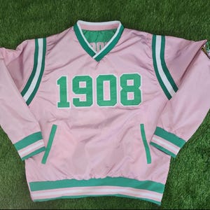 Puede incluir: Chaqueta cortavientos rosa con detalles verdes y blancos. La chaqueta presenta el número "1908" en verde en la parte delantera. Rayas verdes y blancas adornan las mangas y los puños. El cuello y el forro son verdes.