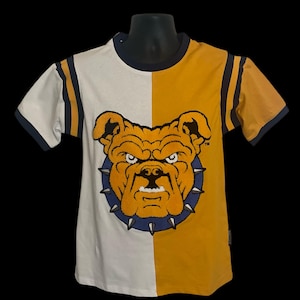May include: A two-toned t-shirt with a bulldog graphic. The shirt is split vertically, with a white left side and a gold right side. The bulldog is gold with a blue spiked collar. The sleeves have blue and gold stripes.