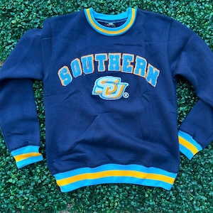 May include: Navy blue sweatshirt with the word "SOUTHERN" arched across the chest in light blue and orange lettering. The sweatshirt has a matching logo below the word "SOUTHERN". The cuffs, collar, and waistline have light blue and orange stripes.