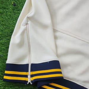 May include: Cream-colored athletic jacket with navy blue and gold stripes. The jacket features a full zipper and navy blue and gold striped cuffs and trim. The jacket is made of a soft, comfortable material.