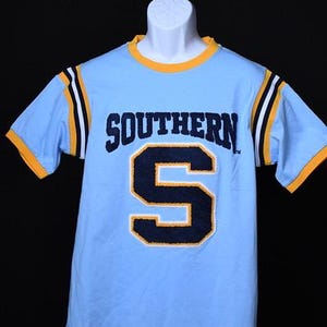 May include: Light blue t-shirt with the word "SOUTHERN" and a large "S" in navy blue and gold. The shirt has gold trim on the collar and sleeves, with navy and white stripes on the sleeves.