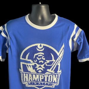 May include: Blue t-shirt with white stripes on the sleeves and collar. The shirt features a white embroidered logo of a pirate with a sword and the text "Hampton Pirates". The shirt is displayed on a black mannequin.