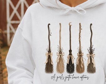 May include: A white hooded sweatshirt featuring a graphic print of five distinct broomsticks arranged in a row, each tied with a small bow. Below the brooms, the text reads A girl's gotta have options in a cursive script font.