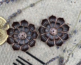 May include: Two copper-colored incense holders shaped like lotus flowers with intricate details. The holders are on a gray concrete surface with black incense sticks and dried lavender sprigs.