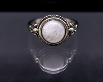 May include: A silver ring with a large, round, white gemstone. The gemstone is set in a gold-colored bezel, surrounded by a twisted rope design. The ring has decorative silver accents on the sides.