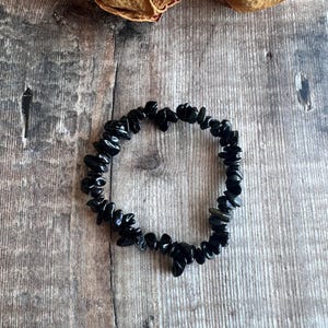May include: A black gemstone bracelet composed of small, irregularly shaped stones. The bracelet is displayed on a wooden surface, with dried flowers in the background. The stones appear to be polished and reflect light, creating a subtle shine.