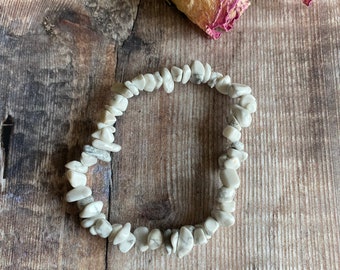 May include: A white howlite chip bracelet with a natural stone look. The bracelet is made of small, irregular shaped chips of white howlite with gray veining. The bracelet is laid out on a wooden surface.