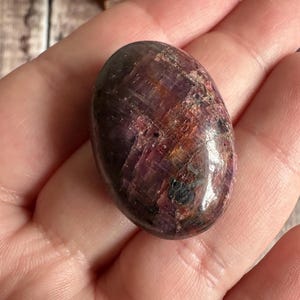 May include: A polished, oval gemstone displaying a blend of deep red, purple, and brown tones. The stone has a smooth, glossy finish, showcasing natural colour and texture variations. This gemstone is a unique piece for jewellery or collection.