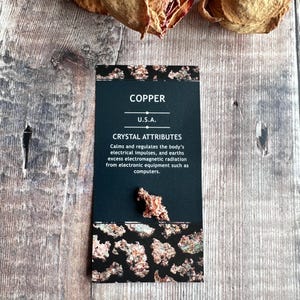 May include: A black card with white text that reads "COPPER U.S.A. CRYSTAL ATTRIBUTES Calms and regulates the body's electrical impulses, and earths excess electromagnetic radiation from electronic equipment such as computers." The card has a copper-coloured border and a small piece of copper ore on the bottom.