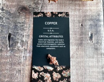 May include: A black card with white text that reads "COPPER U.S.A. CRYSTAL ATTRIBUTES Calms and regulates the body's electrical impulses, and earths excess electromagnetic radiation from electronic equipment such as computers." The card has a copper-colored border and a small piece of copper ore on the bottom.