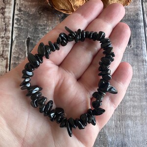 May include: A black gemstone bracelet composed of irregularly shaped, polished black stones. The bracelet is strung together, forming a circular shape. The bracelet is held in a person's hand, with a wooden background.